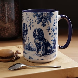May include: A white ceramic mug with a blue and white floral pattern featuring two Cavalier King Charles Spaniels. The mug has a navy blue rim and handle.