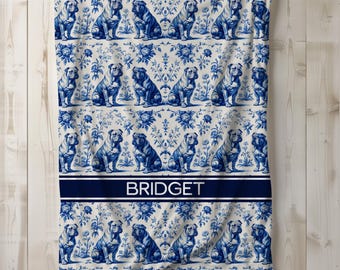 May include: A white and blue patterned blanket featuring a repeating design of bulldog illustrations. The blanket has a navy blue band with the name "BRIDGET" in white text. The design includes floral elements.