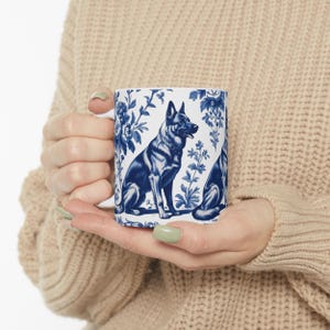 May include: A white ceramic mug with a blue and white floral pattern featuring a German Shepherd dog. The dog is sitting and looking to the right. The mug has a handle on the left side.