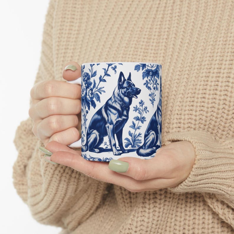 May include: A white ceramic mug with a blue and white floral pattern featuring a German Shepherd dog. The dog is sitting and looking to the right. The mug has a handle on the left side.