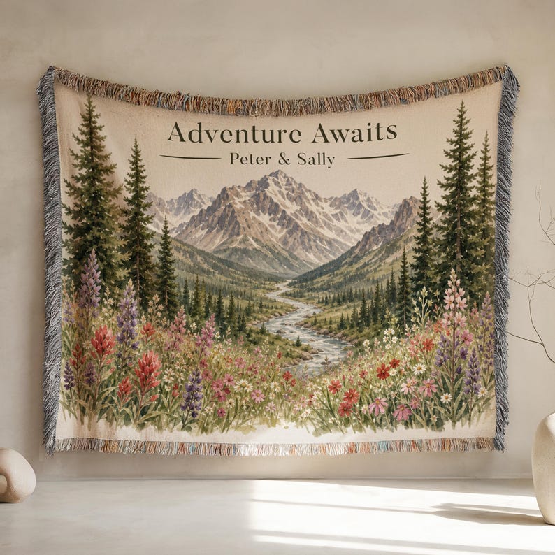 May include: A woven throw blanket featuring a mountain landscape with a winding river, pine trees, and colorful wildflowers. The text "Adventure Awaits" and "Peter & Sally" is printed at the top. The blanket has a fringed edge on all sides.