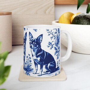 May include: White ceramic mug with a blue and white Chihuahua dog illustration and floral patterns. The mug has a handle and is on a square coaster. The design is reminiscent of traditional blue and white china.
