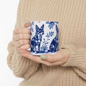 May include: White ceramic mug with a blue and white design of two Chihuahuas and floral patterns. The style is similar to traditional blue and white china. The mug is held by a person wearing a beige jumper.