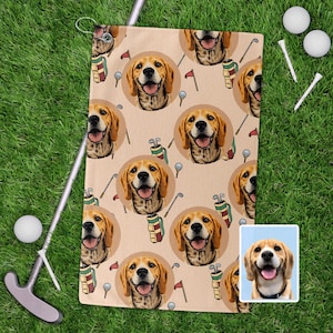 May include: Beige golf towel featuring a repeating pattern of a smiling beagle dog's face, golf clubs, golf balls, and golf flags. The towel is lying on green grass with golf accessories.