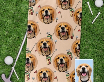 Op de afbeelding: Beige golfhanddoek met een herhaald patroon van een lachende beagle hond, golfclubs, golfballen en vlaggen. De handdoek ligt op groen gras met golfaccessoires.