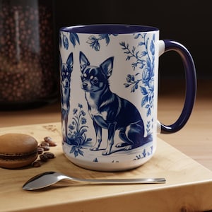 May include: White ceramic mug featuring a blue and white floral and Chihuahua dog design. The mug has a blue handle and rim. A spoon and a pastry are on a wooden surface in front of the mug.