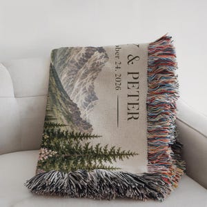 May include: A woven throw blanket featuring a mountain and forest landscape with the text & PETER and October 24, 2026. The blanket has a light beige background, dark green trees, grey mountains, and multicolored fringe along the edges.