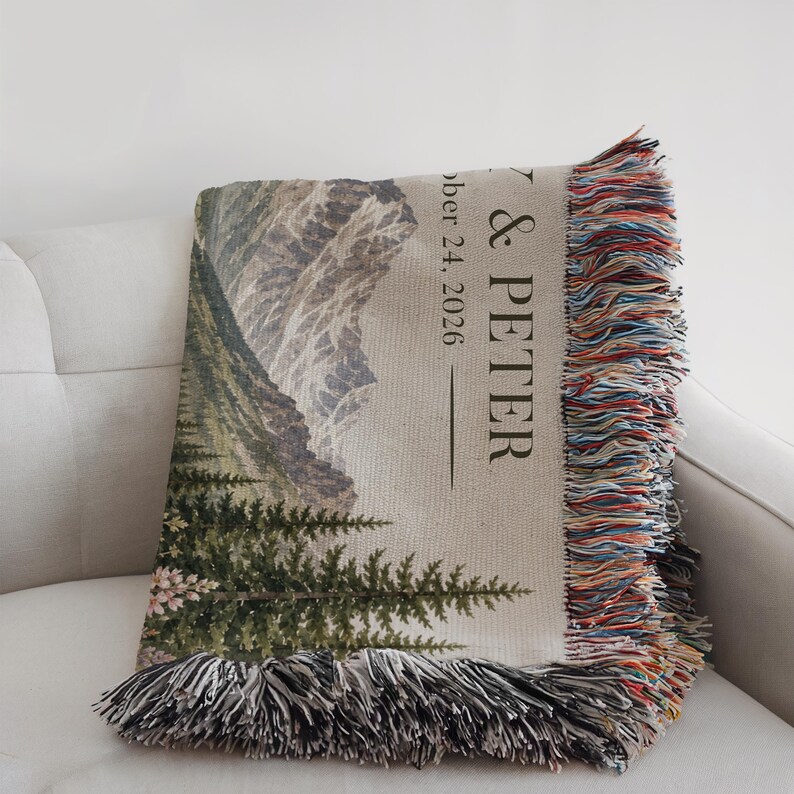 May include: A woven throw blanket featuring a mountain and forest landscape with the text & PETER and October 24, 2026. The blanket has a light beige background, dark green trees, grey mountains, and multicolored fringe along the edges.