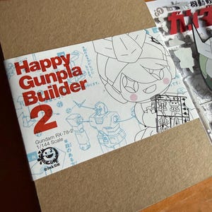 May include: A brown cardboard box with a white label featuring the text "Happy Gunpla Builder 2". The label includes illustrations of a Gundam RX-78-2 model and a cartoon character. The scale is 1/144.