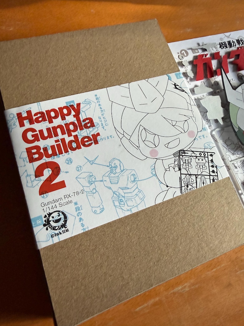 May include: A brown cardboard box with a white label featuring the text "Happy Gunpla Builder 2". The label includes illustrations of a Gundam RX-78-2 model and a cartoon character. The scale is 1/144.