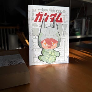 May include: A white and red Japanese-language book featuring a cartoon character in a green and gray helmet holding a plastic model kit. The word "ガンダム" is prominently displayed in red. The book is on a wooden table.