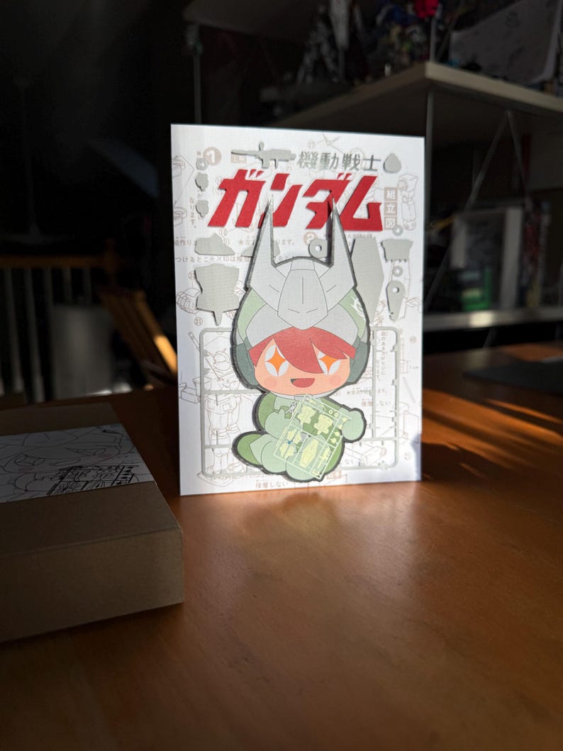 May include: A white and red Japanese-language book featuring a cartoon character in a green and gray helmet holding a plastic model kit. The word "ガンダム" is prominently displayed in red. The book is on a wooden table.
