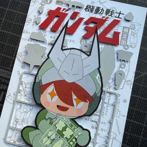 May include: A colorful papercraft featuring a cartoon character in a green and white military-style outfit, holding a clear plastic sheet with small parts. The background includes Japanese text and red lettering that says "Gundam".