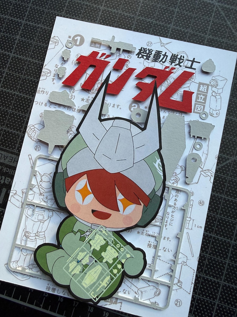 May include: A colorful papercraft featuring a cartoon character in a green and white military-style outfit, holding a clear plastic sheet with small parts. The background includes Japanese text and red lettering that says "Gundam".