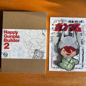 May include: A brown cardboard box with a white and red label that reads "Happy Gunpla Builder 2". Next to it is a white card with a cartoon character and Japanese text. The character is wearing a helmet and holding a small clear plastic sheet.