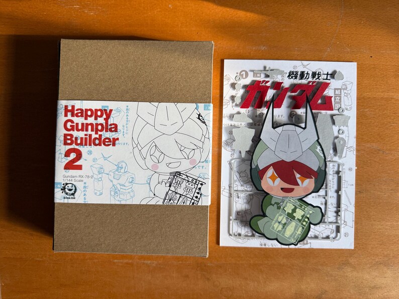 May include: A brown cardboard box with a white and red label that reads "Happy Gunpla Builder 2". Next to it is a white card with a cartoon character and Japanese text. The character is wearing a helmet and holding a small clear plastic sheet.