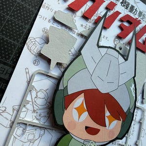 May include: A colorful paper cutout of a cartoon character wearing a helmet, with orange star-shaped eyes and a smiling expression. The character is holding a clear plastic sheet with small parts. Japanese text and cut-out pieces are visible.