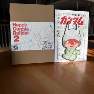 May include: A brown cardboard box and a white booklet with Japanese text and illustrations. The box reads "Happy Gunpla Builder 2" with a cartoon character. The booklet features a Gundam character and the text "機動戦士ガンダム".