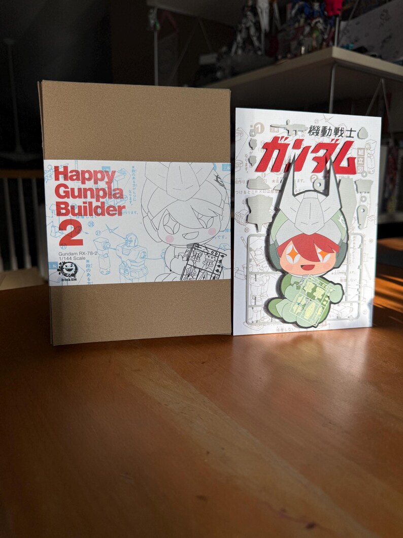 May include: A brown cardboard box and a white booklet with Japanese text and illustrations. The box reads "Happy Gunpla Builder 2" with a cartoon character. The booklet features a Gundam character and the text "機動戦士ガンダム".