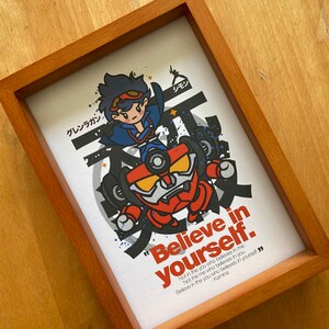 K&ouml;nnte beinhalten: Ein gerahmter Druck mit einer Cartoon-Illustration eines Charakters in einem roten und gelben Roboteranzug. Der Druck zeigt den Text "Glaube an dich selbst." und ein Zitat von Kamina aus der Anime-Serie Gurren Lagann.
