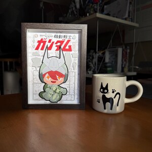 May include: A framed illustration featuring a cartoon character with red hair and a green outfit, with Japanese text. Beside it is a white mug with a black cat design.