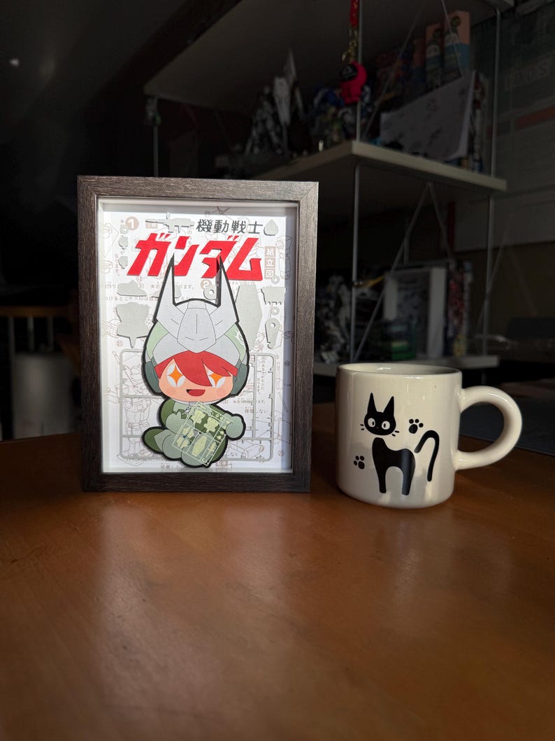 May include: A framed illustration featuring a cartoon character with red hair and a green outfit, with Japanese text. Beside it is a white mug with a black cat design.