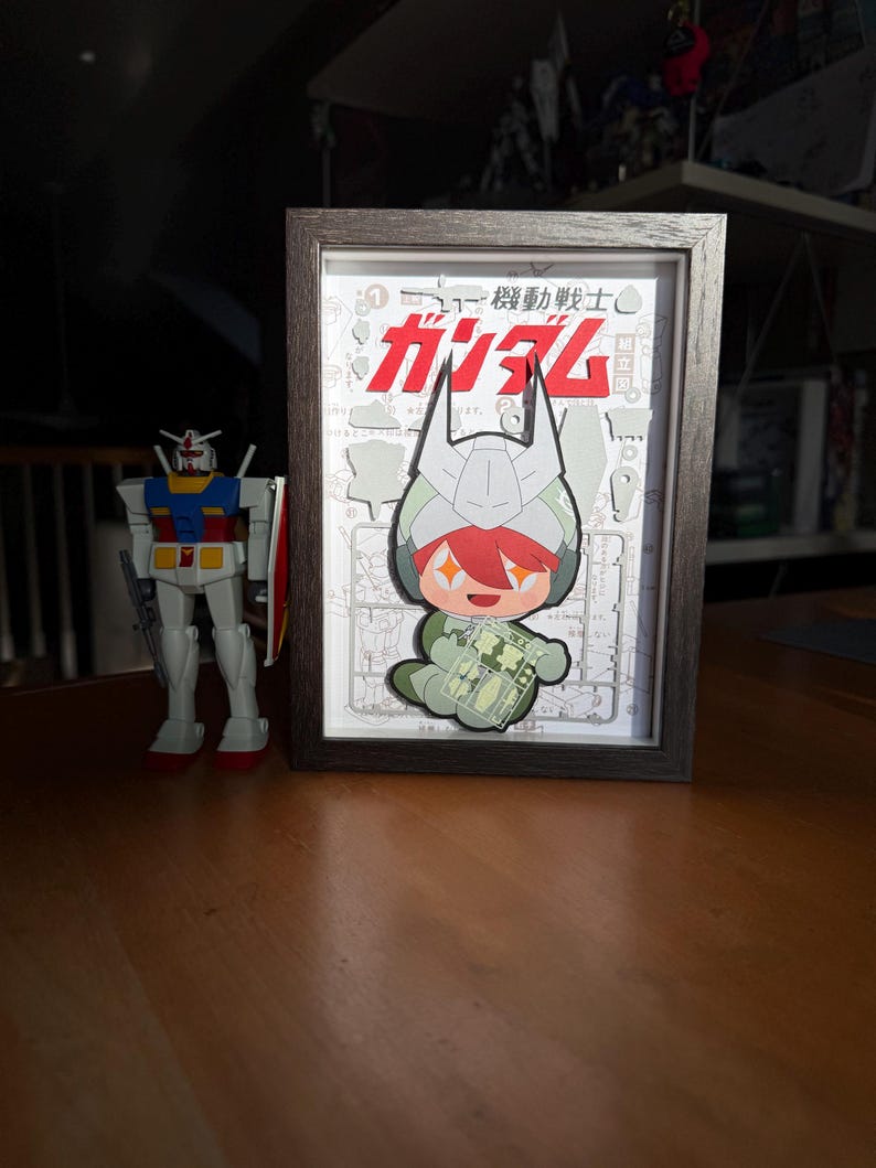 May include: A framed artwork featuring a cartoon character with red hair and a green outfit, set against a white background with Japanese text. A Gundam model stands to the left. The frame is dark brown, and the overall theme is anime.