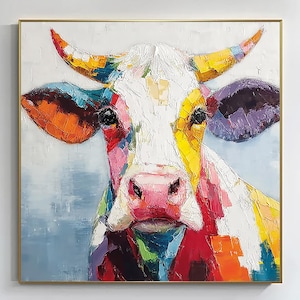 May include: A colorful cow portrait painting with a gold frame. The cow's face is painted with vibrant hues of red, orange, yellow, blue, and purple. The artwork is displayed on a white wall.