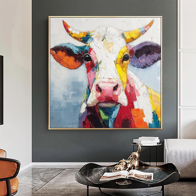 May include: A colorful cow portrait painting with a gold frame. The cow's face is painted with bold brushstrokes in a variety of colors, including red, yellow, orange, and purple. The background is a soft blue. The artwork is a modern, abstract style.