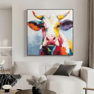 May include: A colorful cow portrait painting with a black frame. The cow's face is depicted with vibrant brushstrokes of red, yellow, orange, purple, and white. The artwork is displayed in a living room setting, above a white sofa.