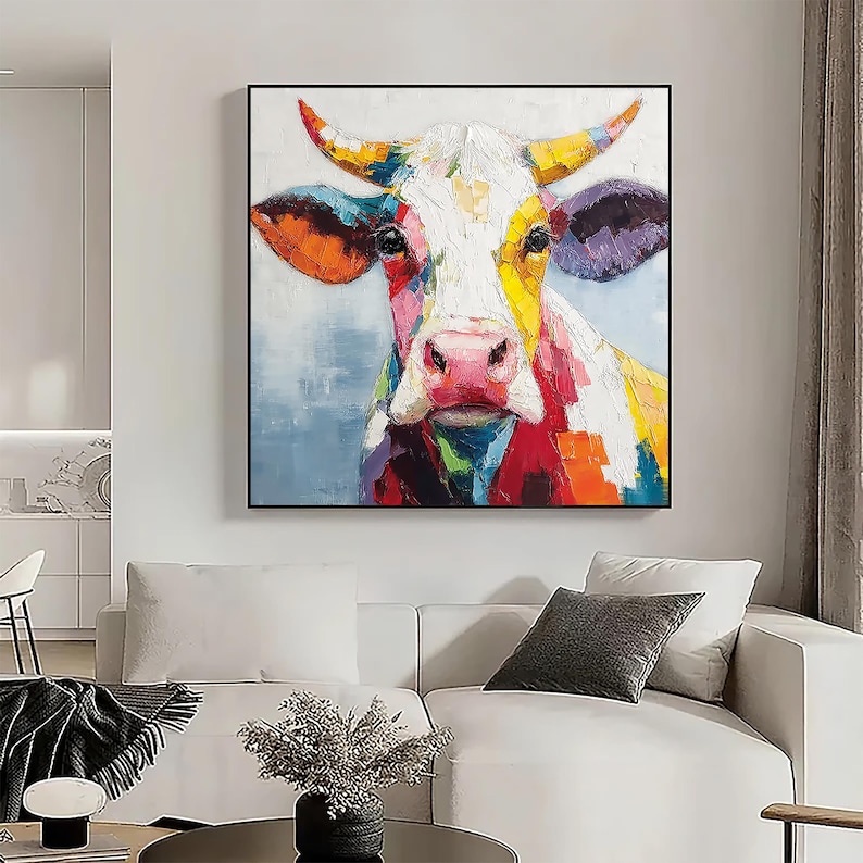 May include: A colorful cow portrait painting with a black frame. The cow's face is depicted with vibrant brushstrokes of red, yellow, orange, purple, and white. The artwork is displayed in a living room setting, above a white sofa.