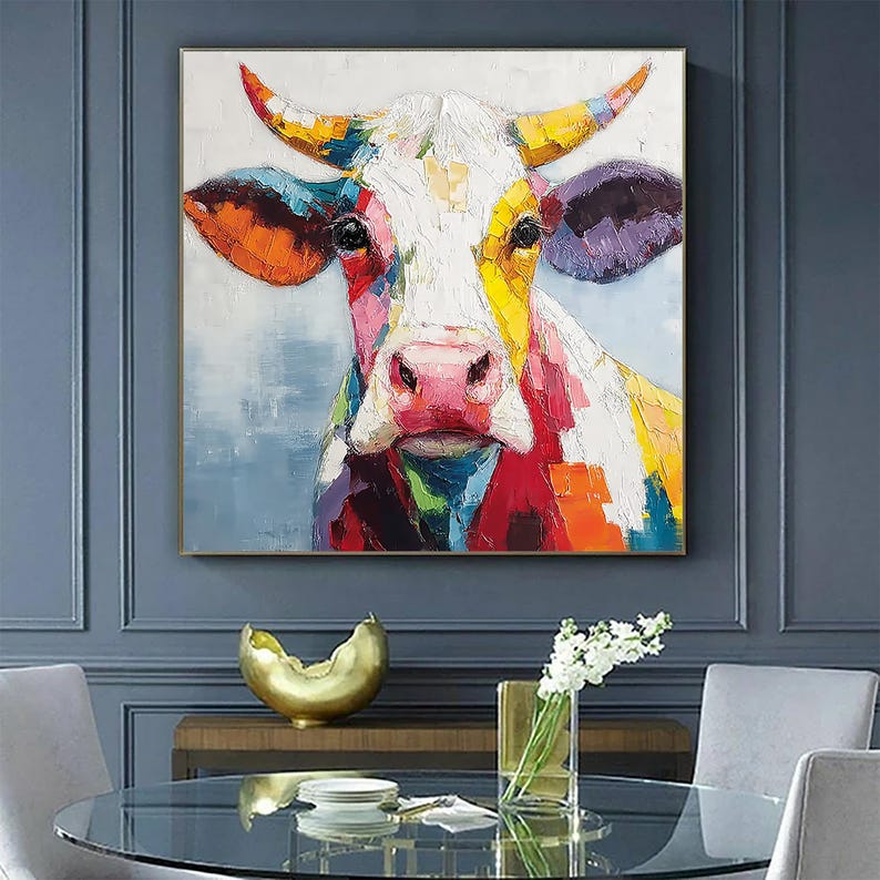 May include: A colorful oil painting of a cow's face. The cow is depicted with a white face, a pink nose, and multi-colored accents of red, orange, yellow, blue, and purple. The artwork is framed in a gold-colored frame.