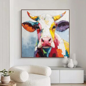 May include: A colorful cow painting with a square frame. The cow's face is white with a pink nose, and its horns are yellow and orange. The painting uses a palette of bright colors, including red, blue, and green.