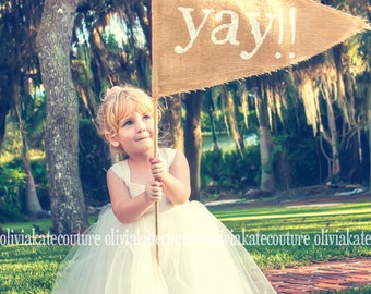May include: A young girl wearing a white tulle dress holds a burlap banner with the word "yay!" written in white paint. The banner is triangular and has frayed edges. The girl is smiling and looking up at the banner.