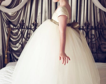 May include: A young girl wearing a white tulle dress with a sage green sash and a flower in her hair. She is standing on a white bedspread.
