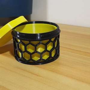 May include: A black and yellow cylindrical container with a honeycomb pattern. The container has a removable lid.