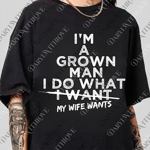 May include: Black t-shirt with white text that reads "I'M A GROWN MAN I DO WHAT I WANT" with a line through "I WANT" and "MY WIFE WANTS" below. The shirt is a casual style.