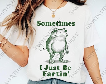 May include: White t-shirt with a green illustration of a frog and the text "Sometimes I Just Be Fartin'". The frog is depicted with a comical expression, and a puff of gas is drawn next to it. The text is in a playful, casual font.