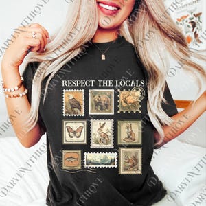 A woman with long blonde hair is smiling and posing with a black t-shirt featuring a graphic design of various animals and postage stamps.