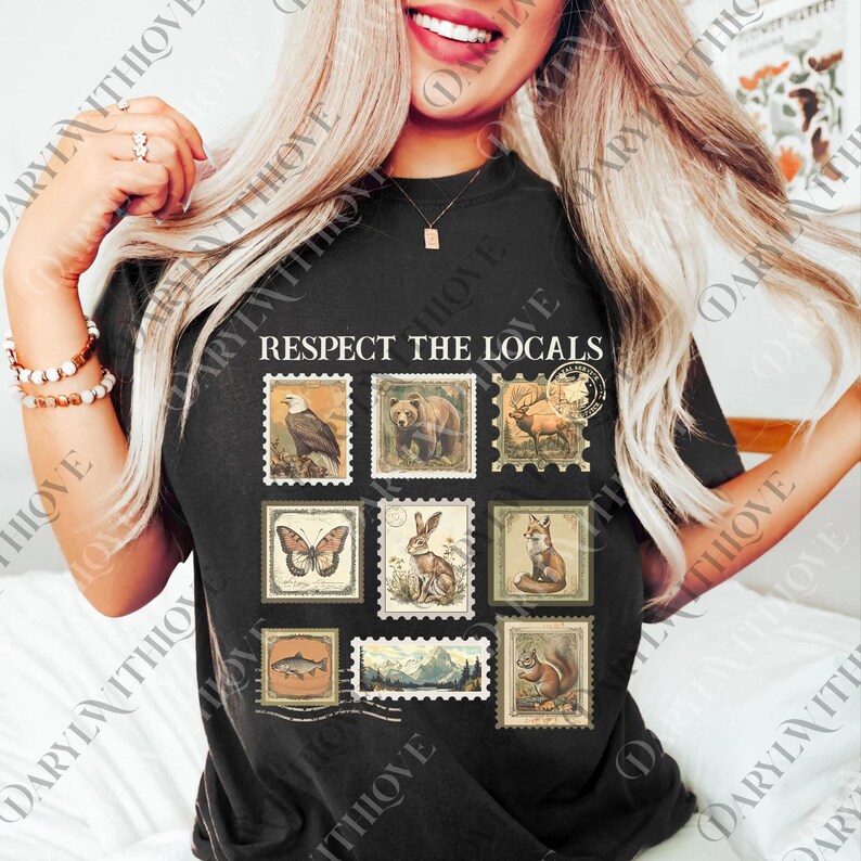 A woman with long blonde hair is smiling and posing with a black t-shirt featuring a graphic design of various animals and postage stamps.