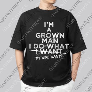 May include: Black t-shirt with white text that reads "I'M A GROWN MAN I DO WHAT MY WIFE WANTS." The shirt is a crew neck and has short sleeves. The text is in a bold, sans-serif font.