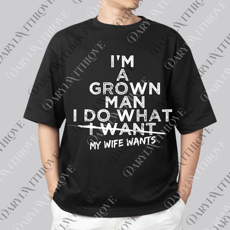 May include: Black t-shirt with white text that reads "I'M A GROWN MAN I DO WHAT MY WIFE WANTS." The shirt is a crew neck and has short sleeves. The text is in a bold, sans-serif font.