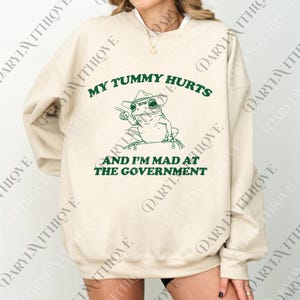 May include: Cream-colored sweatshirt with a green graphic of a frog wearing a cowboy hat and pointing. The text reads "MY TUMMY HURTS AND I'M MAD AT THE GOVERNMENT". A casual, humorous design.