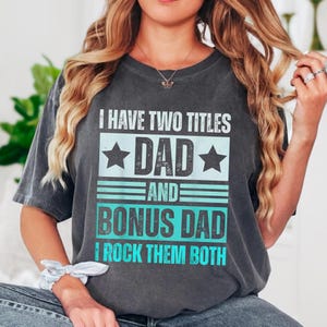 Può includere: Una t-shirt grigio antracite a maniche corte con il testo bianco e turchese I HAVE TWO TITLES DAD AND BONUS DAD I ROCK THEM BOTH, accentuato da due stelle e linee orizzontali, stampato sul davanti in uno stile dall'aspetto vissuto.