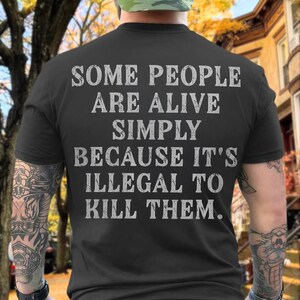 Könnte beinhalten: Ein schwarzes Kurzarm-T-Shirt mit dem weißen Text im Used-Look: SOME PEOPLE ARE ALIVE SIMPLY BECAUSE IT'S ILLEGAL TO KILL THEM. Das T-Shirt hat einen Rundhalsausschnitt und wird von hinten gezeigt, wobei die gedruckte Botschaft deutlich sichtbar ist.