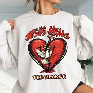May include: A white crewneck sweatshirt featuring a graphic print of a large red heart with a cross and a white dove. The text "JESUS HEALS" is arched above the heart, and "THE BROKEN" is printed below in a bold, red, and yellow font style.