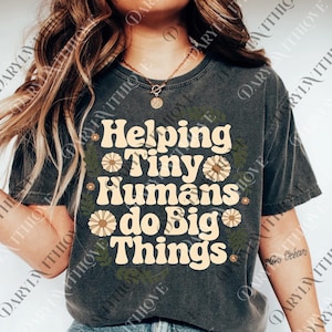 May include: Dark gray t-shirt with the text "Helping Tiny Humans do Big Things" in a retro font. The design includes daisy and leaf accents. A casual, comfortable tee.