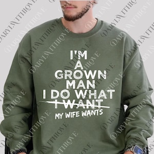 May include: Olive green sweatshirt with white text that reads "I'M A GROWN MAN I DO WHAT MY WIFE WANTS". The sweatshirt has a crew neck and long sleeves. The text is centered on the front of the shirt.