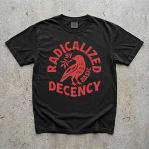 Pode incluir: Camiseta preta com um gráfico vermelho desgastado. O gráfico apresenta um pássaro segurando um galho e as palavras "RADICALIZED BY BASIC DECENCY" em um design circular. A camiseta tem gola redonda e mangas curtas.