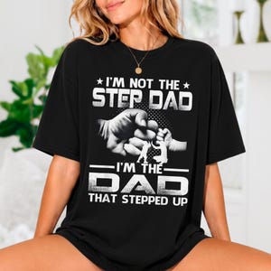 May include: A black short-sleeve t-shirt featuring white graphic text that reads I'M NOT THE STEP DAD I'M THE DAD THAT STEPPED UP. The design includes a central illustration of an adult fist bumping a child's fist with a dotted background pattern.